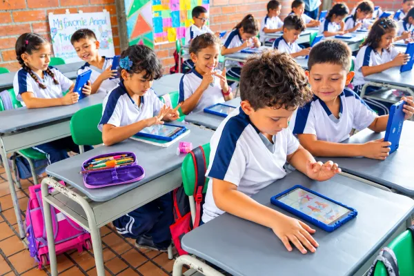 Estudiantes de primaria con uniforme trabajan concentrados en sus tabletas, cada uno en su pupitre, en un aula con muro de ladrillo