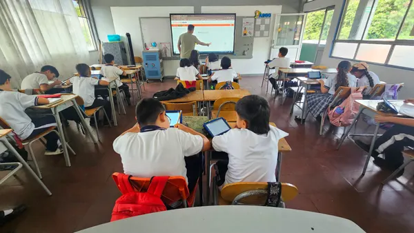Un docente abre la plataforma eClass en la pantalla interactiva mientras sus estudiantes trabajan con tabletas y cuadernos