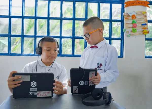 Dos estudiantes de primaria con uniforme trabajan cada uno en su tableta AVACOM, uno de ellos con audífonos, en un aula con ventanal de bloques de vidrio
