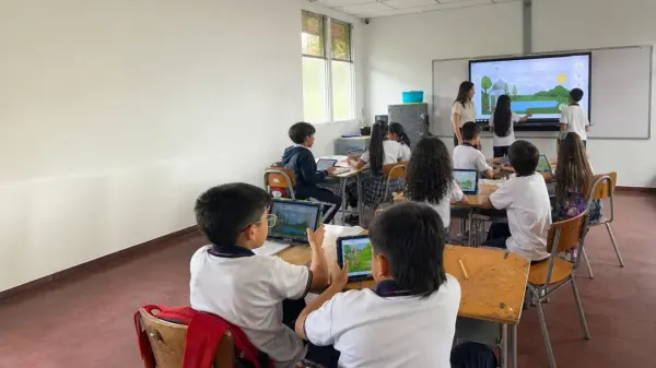 Una estudiante trabaja en la pantalla interactiva con un paisaje del entorno mientras la docente la acompaña y la clase sigue desde sus tabletas